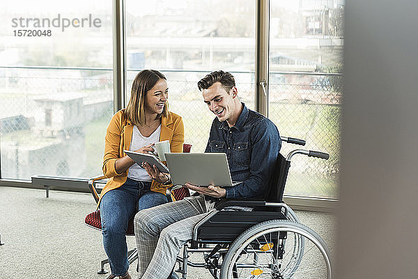 Glücklicher junger Geschäftsmann mit Laptop im Rollstuhl und Geschäftsfrau mit Tablett im Büro