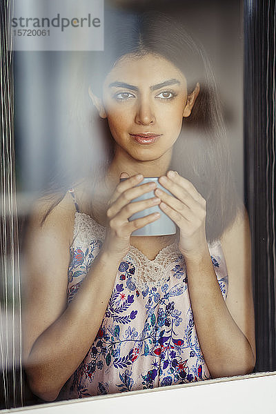 Porträt einer jungen Frau mit Kaffeebecher hinter Fensterscheibe