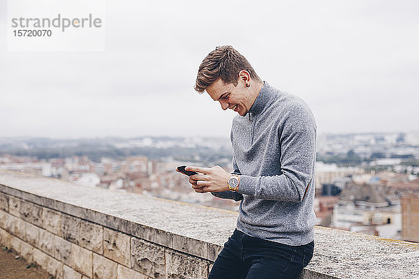 Blonder junger Mann mit einem Pullover  der in Lleida lächelnd eine Nachricht auf sein Handy schreibt