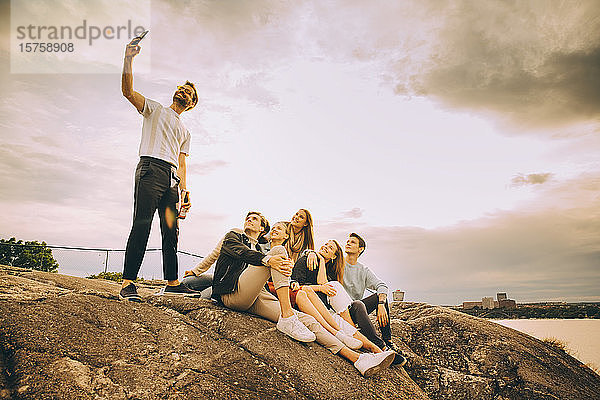 Mann nimmt Selfie mit Freunden auf Felsformation gegen den Himmel beim Picknick bei Sonnenuntergang