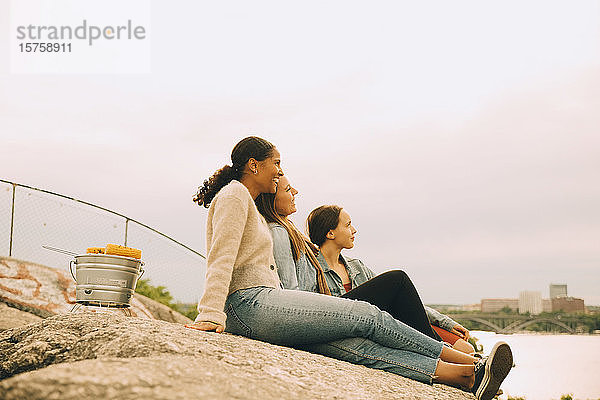 Glückliche Freundinnen schauen weg  während sie auf einer Felsformation am Seeufer gegen den Himmel sitzen
