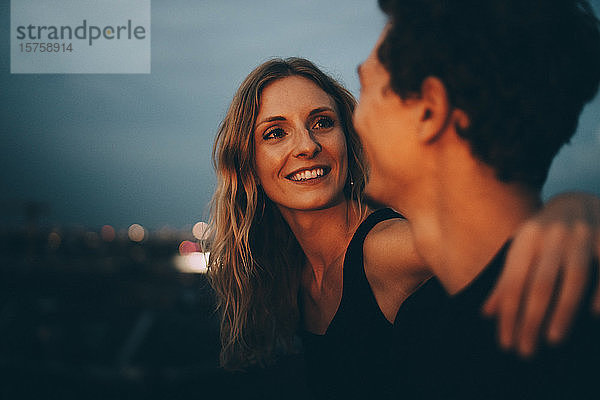 Lächelnde Frau mit umarmtem Arm  die den Mann ansieht  während sie in der Abenddämmerung auf der Terrasse sitzt