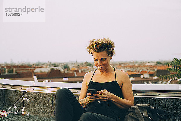 Lächelnde Frau mittleren Alters  die ein Mobiltelefon benutzt  während sie auf einer Terrasse gegen den Himmel in der Stadt sitzt