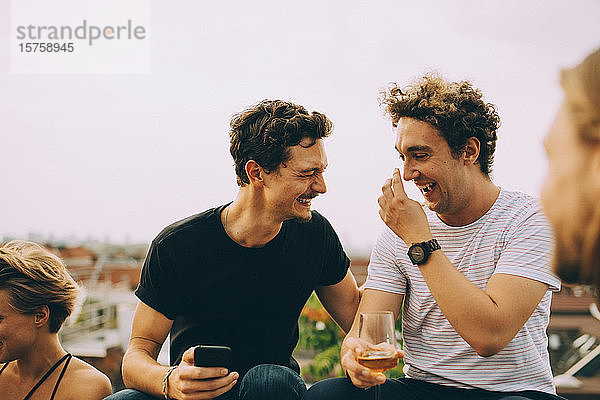 Glücklicher Mann zeigt einem Freund sein Handy und trinkt während der Party auf der Terrasse