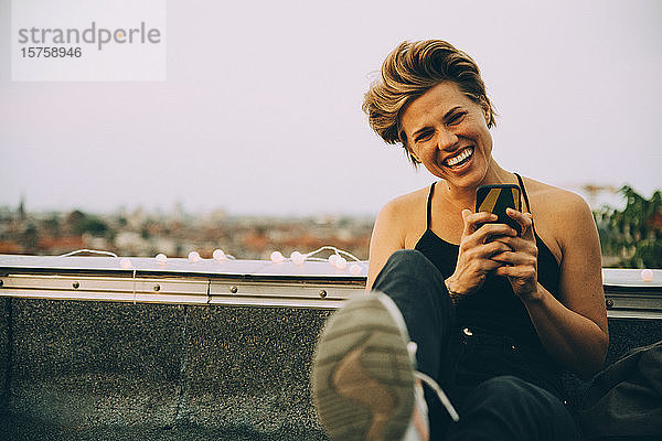 Porträt einer fröhlichen Frau  die ein Mobiltelefon benutzt  während sie auf einer Terrasse gegen den Himmel sitzt