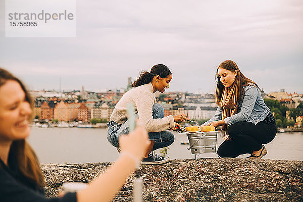 Freundinnen rösten beim Picknick Zuckermais auf einer Felsformation am See