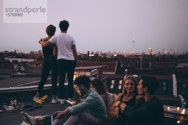 Ein Paar betrachtet die Stadt  während Freunde während einer Dachparty auf der Terrasse entspannen