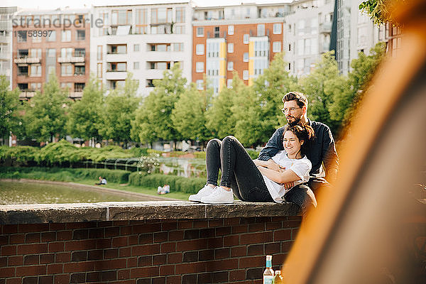 Junger Mann mit lächelnder Partnerin schaut weg  während er auf einer Stützmauer sitzt