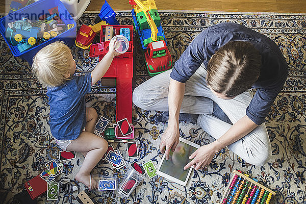 Hochwinkelansicht eines blonden kleinen Jungen  der zu Hause im Wohnzimmer mit einem digitalen Tablett Spielzeug des Vaters spielt