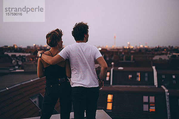 Rückansicht eines Paares  das mit dem Arm um die Stadt blickt  während es in der Dämmerung auf der Terrasse steht