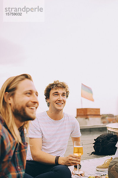 Lächelnde männliche Freunde genießen Bier  während sie während einer Party gegen den Himmel auf die Terrasse schauen