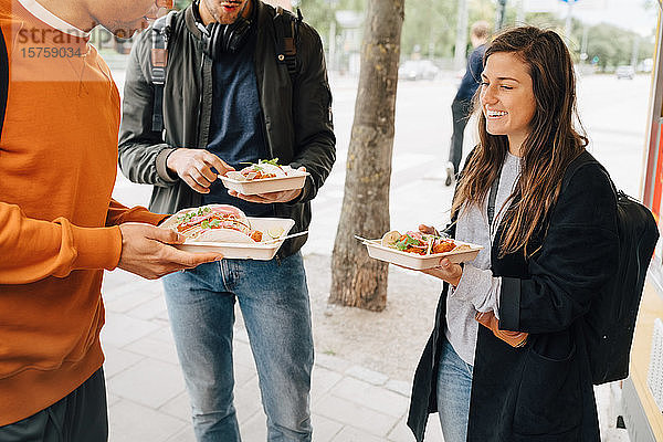 Lächelnde junge Frau genießt Essen mit männlichen Freunden  während sie mit einem Speisewagen auf der Straße steht