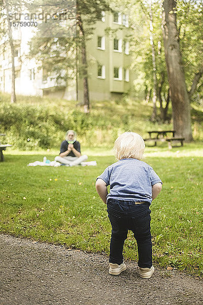 Rückansicht in voller Länge eines auf einem Fußweg stehenden Jungen mit einem im Park sitzenden Vater