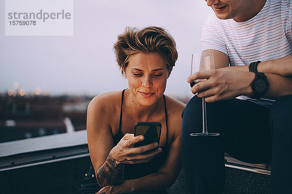 Frau benutzt Smartphone  während sie bei einem Freund sitzt und Wein auf der Terrasse trinkt