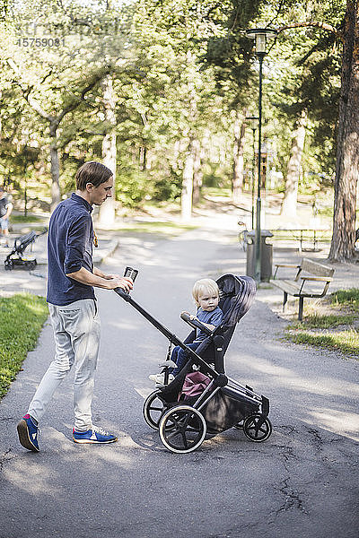 Mann läuft in voller Länge  während er seinen Sohn im Kinderwagen im öffentlichen Park schiebt