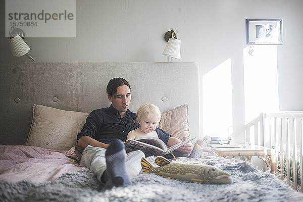 Vater liest mit Kleinkind Sohn Bilderbuch  während er zu Hause auf dem Bett sitzt