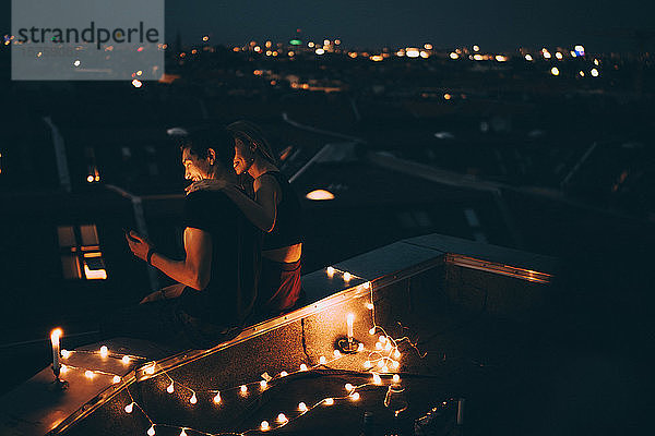 Hochwinkelansicht eines Paares  das in der Abenddämmerung auf einer beleuchteten Terrasse in der Stadt sitzt und auf sein Mobiltelefon schaut