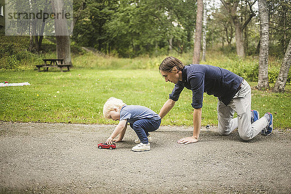 Vater kniet  während der Sohn mit einem Spielzeugauto auf dem Fußweg gegen Pflanzen im Park spielt