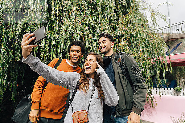 Lächelnde Freunde beim Selbermachen mit dem Handy  während sie in der Stadt am Baum stehen