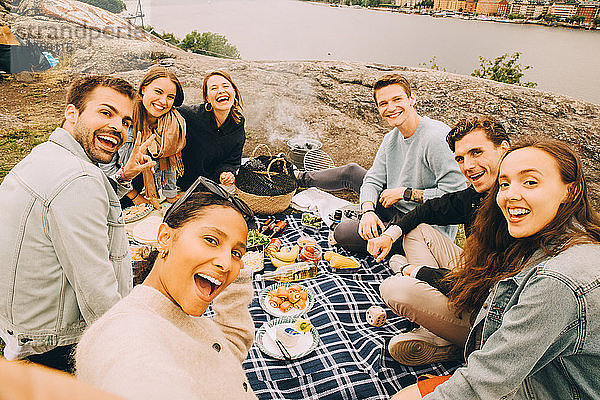 Porträt von fröhlichen Freunden  die im Sommer auf dem Feld am See picknicken