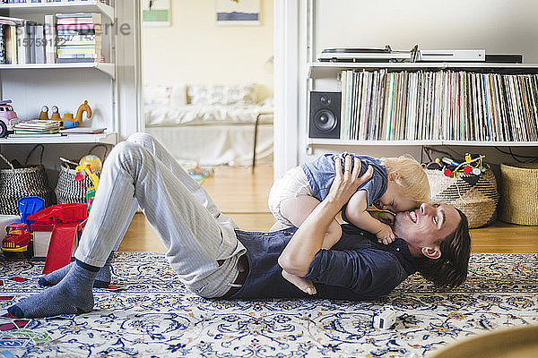 Lächelnder Mann liegt auf dem Teppich  während er zu Hause im Wohnzimmer mit seinem Kleinkind spielt