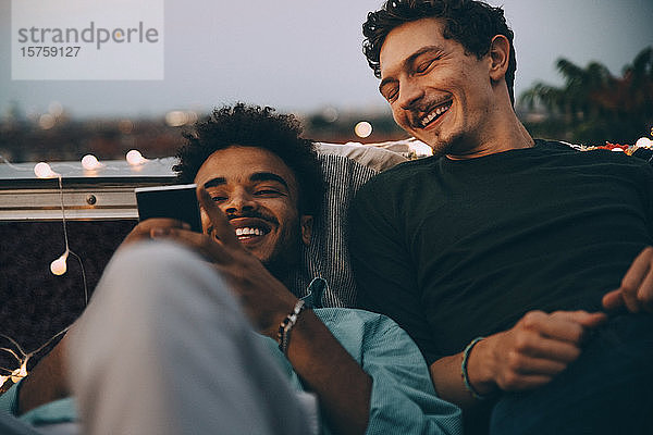 Glückliche männliche Freunde schauen auf Smartphone und entspannen sich auf der Terrasse während einer Dachparty in der Stadt