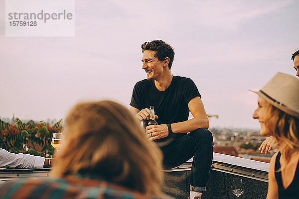 Lächelnder Mann schaut weg  während er mit Freunden Bier auf einer Dachparty gegen den Himmel genießt