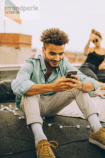 Lächelnder junger Mann benutzt Smartphone  während er bei einer Party auf der Terrasse sitzt