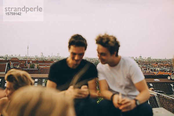 Freunde  die während einer Dachparty auf der Terrasse sitzen und auf ihr Handy schauen