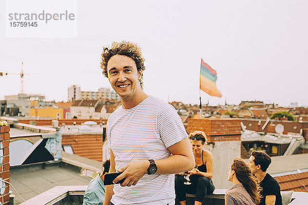 Porträt eines lächelnden jungen Mannes mit Mobiltelefon  während sich Freunde während einer Party auf der Terrasse vergnügen
