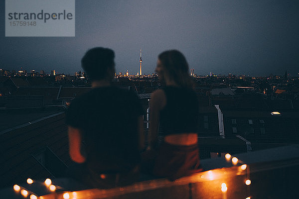 Rückansicht eines Paares  das in der Abenddämmerung auf einer Terrasse gegen das Stadtbild sitzt