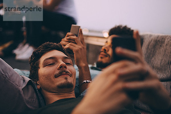 Junge männliche Freunde benutzen Smartphones  während sie in der Dämmerung auf der Terrasse liegen