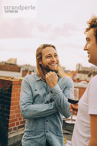 Lächelnde männliche Freunde unterhalten sich im Stehen auf der Terrasse bei einer Dachfeier