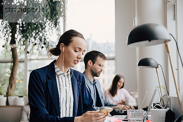 Geschäftsfrau  die Musik über Kopfhörer hört  während sie mit Kollegen am Schreibtisch im Büro sitzt