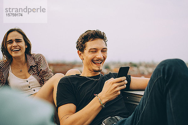 Fröhlicher Mann schaut auf Smartphone  während Freundin Spaß auf Dachparty hat