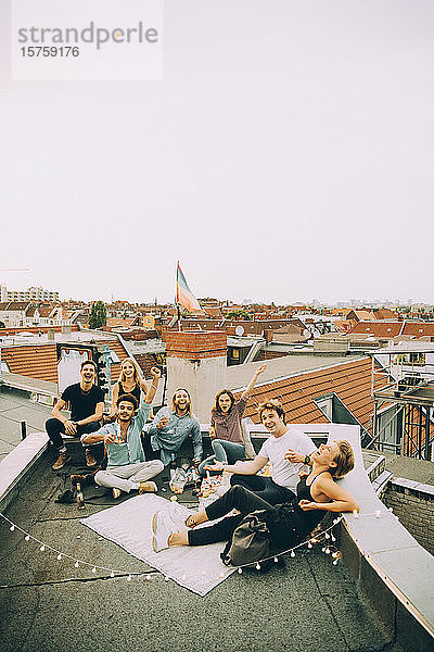 Fröhliche männliche und weibliche Freunde amüsieren sich auf der Terrasse während einer Dachparty in der Stadt gegen den Himmel