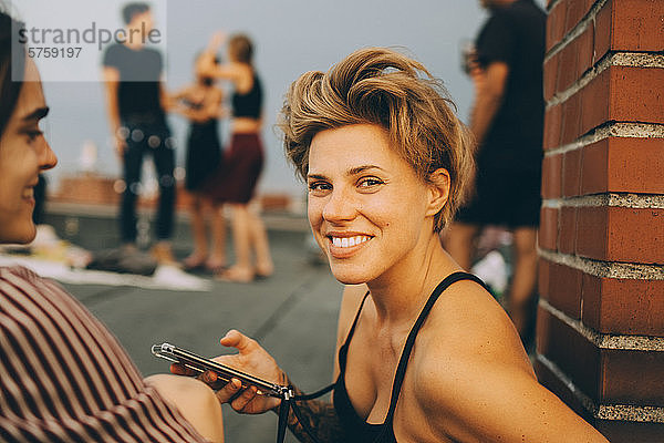 Porträt einer lächelnden Frau mit Smartphone in der Hand  die während einer Dachparty mit einem Freund zusammensitzt