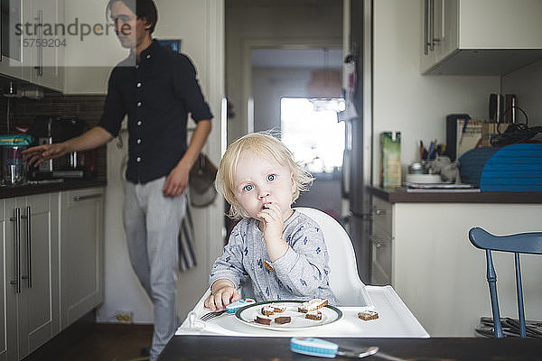 Porträt eines blonden Jungen beim Essen auf einem Hochstuhl in der Küche mit dem Vater im Hintergrund