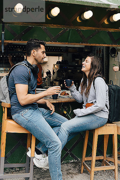 Lächelnde Freunde in voller Länge  die Kaffeetassen in der Hand halten  während sie an einem Imbissstand in der Stadt sitzen