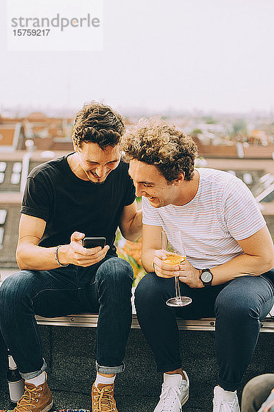 Mann zeigt einem Freund sein Handy und trinkt während der Party auf der Terrasse