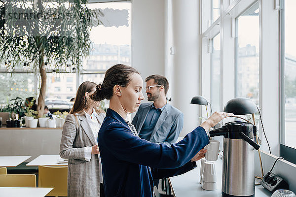 Seitenansicht einer Geschäftsfrau  die Kaffee trinkt  während die Kollegen im Hintergrund diskutieren