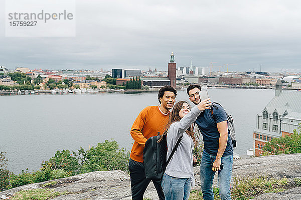 Lächelnde Freunde  die sich mit dem Handy selbstständig machen  während sie am Fluss in der Stadt gegen den Himmel stehen