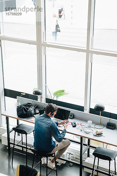 Hochwinkelansicht eines Mannes  der während der Arbeit am Schreibtisch im Büro Musik auf einem Smartphone hört