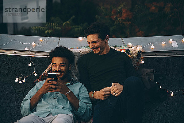 Lächelnde männliche Freunde schauen auf ein Smartphone und sitzen während einer Dachparty in der Stadt auf der Terrasse