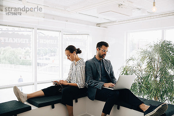 Männliche und weibliche Kollegen nutzen Technologien  während sie im kreativen Büro auf dem Flur sitzen