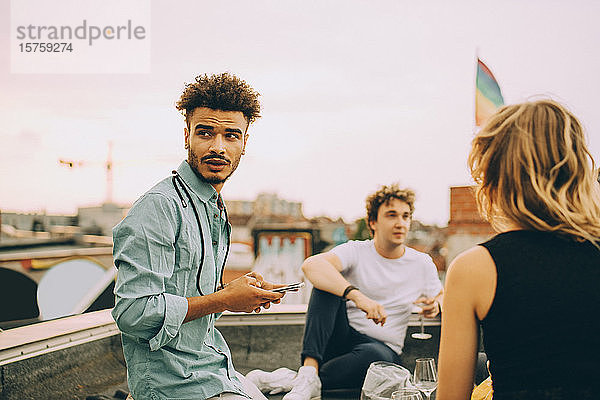 Junger Mann verschickt Textnachrichten auf Smartphone  während er bei einer Dachparty mit Freunden auf der Terrasse sitzt