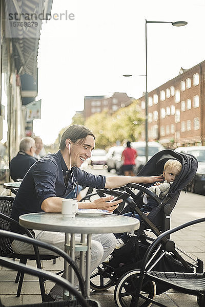 Lächelnder Mann hört Musik über Kopfhörer von einem Smartphone aus  während er mit dem Baby im Kinderwagen im Straßencafé auf dem Bürgersteig sitzt