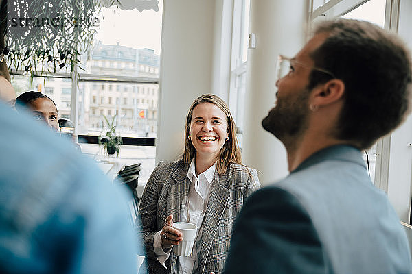 Lächelnde Geschäftsfrau trinkt Kaffee mit Kollegen am kreativen Arbeitsplatz