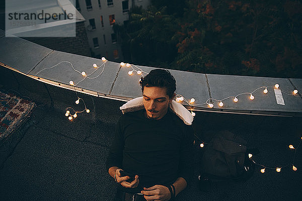Hochwinkelansicht eines jungen Mannes  der ein Smartphone benutzt  während er sich in der Dämmerung auf einer beleuchteten Terrasse entspannt