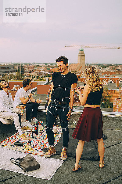 Verspielte Frau bindet dem Mann eine Leuchtschnur um den Hals  während sich Freunde auf der Dachparty amüsieren